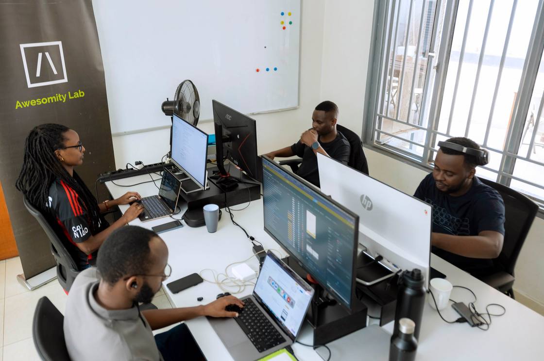 Four Awesomity Lab engineers at their desks in the Kigali office, with monitors, a whiteboard and an Awesomity Lab banner.